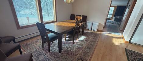 A dining area with a wooden table and chairs, large windows, and a chandelier.