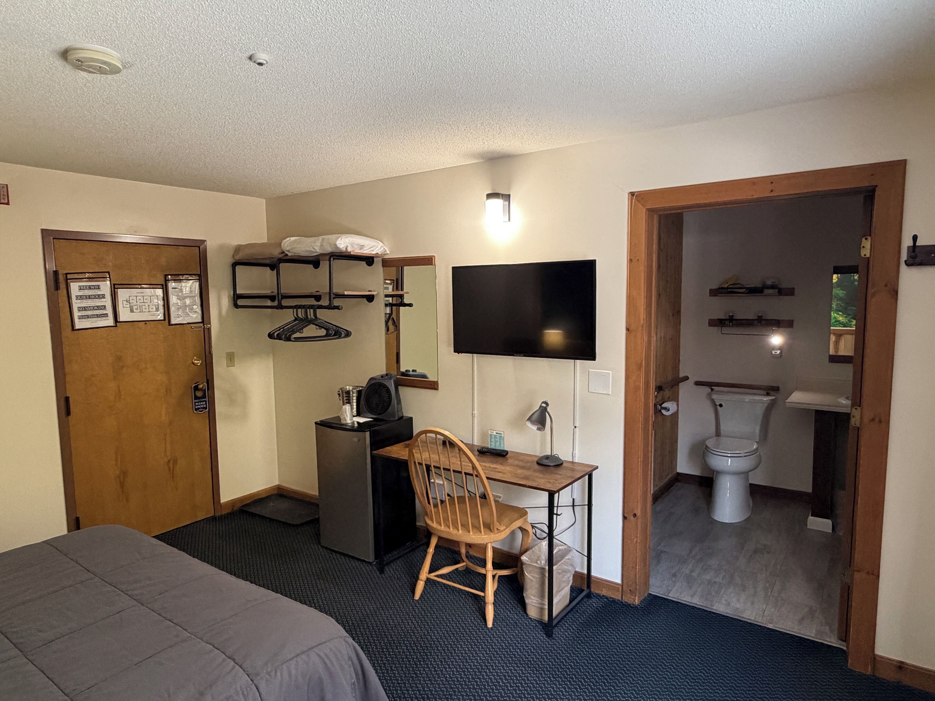 A hotel room with a bed, a desk with a chair, a TV, a small fridge, a heater, and a bathroom with a toilet and sink.