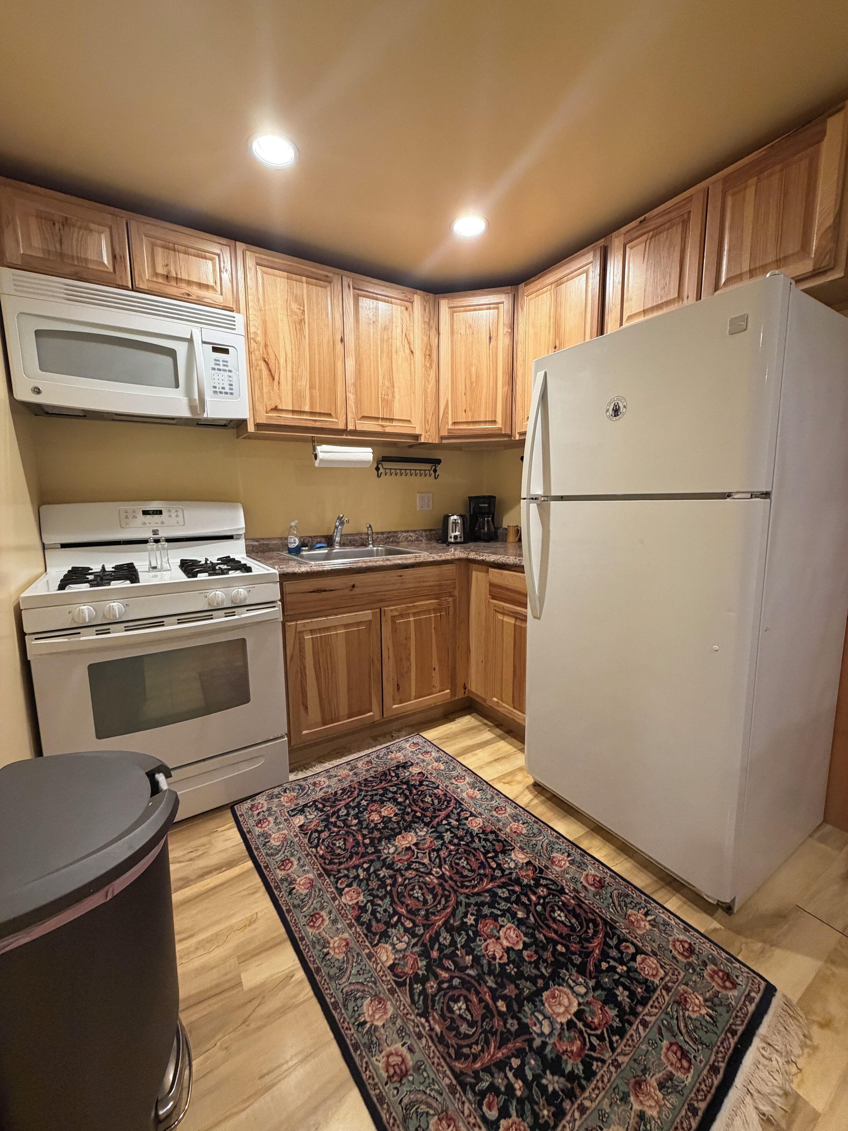 A kitchen with wooden cabinets, a microwave, stove, oven, refrigerator, sink, and a patterned rug.