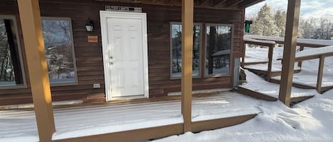 Uma cabana de madeira com uma porta branca identificada como "Outer Limits", um alpendre coberto e um deck coberto de neve.