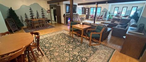 A spacious living room with a wooden dining table, a sofa, and a coffee table. There is a large rug on the floor and a fireplace in the background.