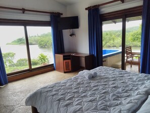 A hotel room with a bed, a desk, a chair, and a TV. The room has large windows with a view of a lake and greenery. - Casa Caletas (Bejuco)