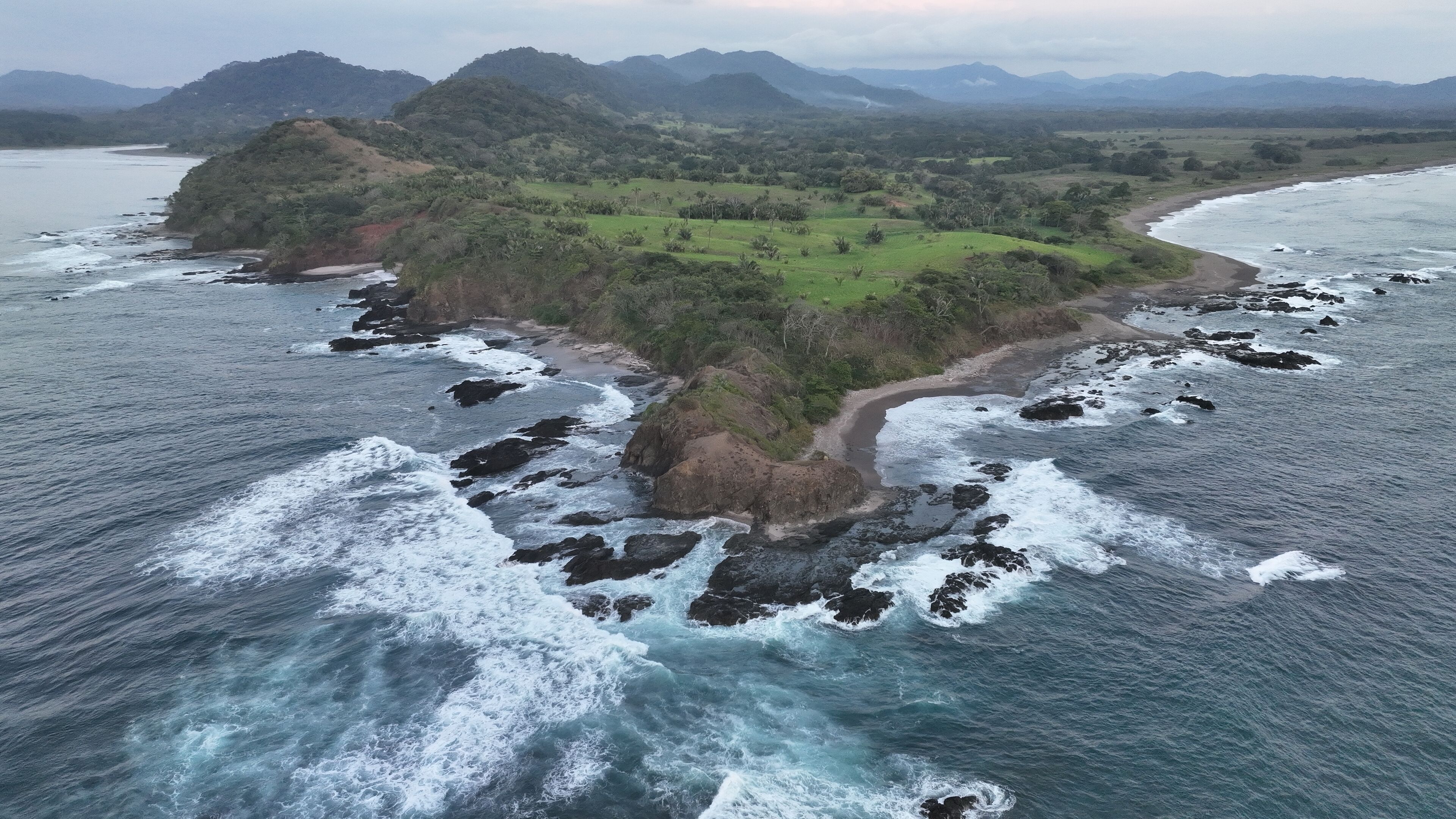 An aerial view of a coastal landscape with a small island featuring a dirt path, surrounded by rocky shores and waves.