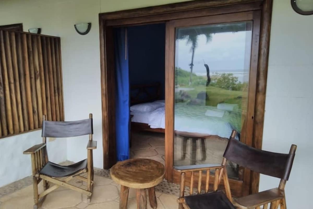 Una habitación con cama, una silla de madera, una mesita redonda y una puerta de cristal que da a un balcón con vistas a la playa.