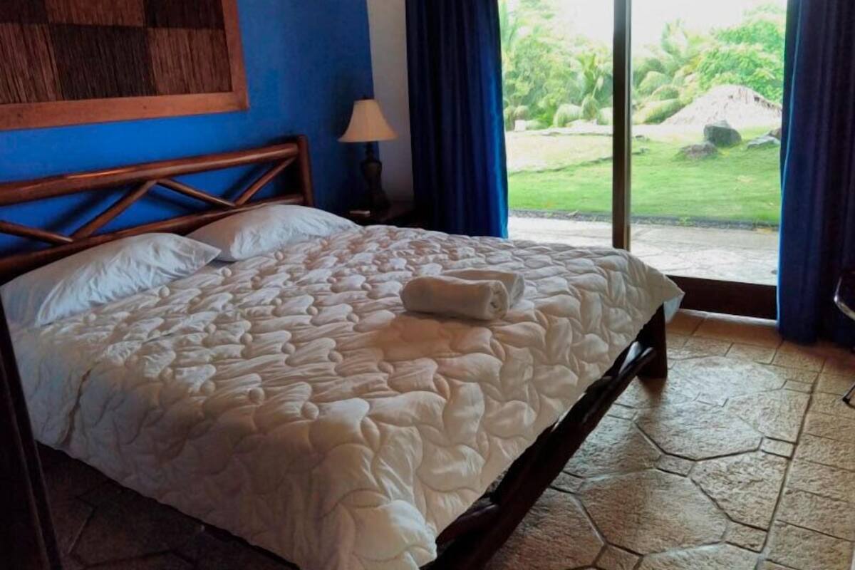 Un dormitorio con una cama grande, cabecera de madera, mesita de noche con lámpara y vista a un jardín a través de una puerta corrediza.
