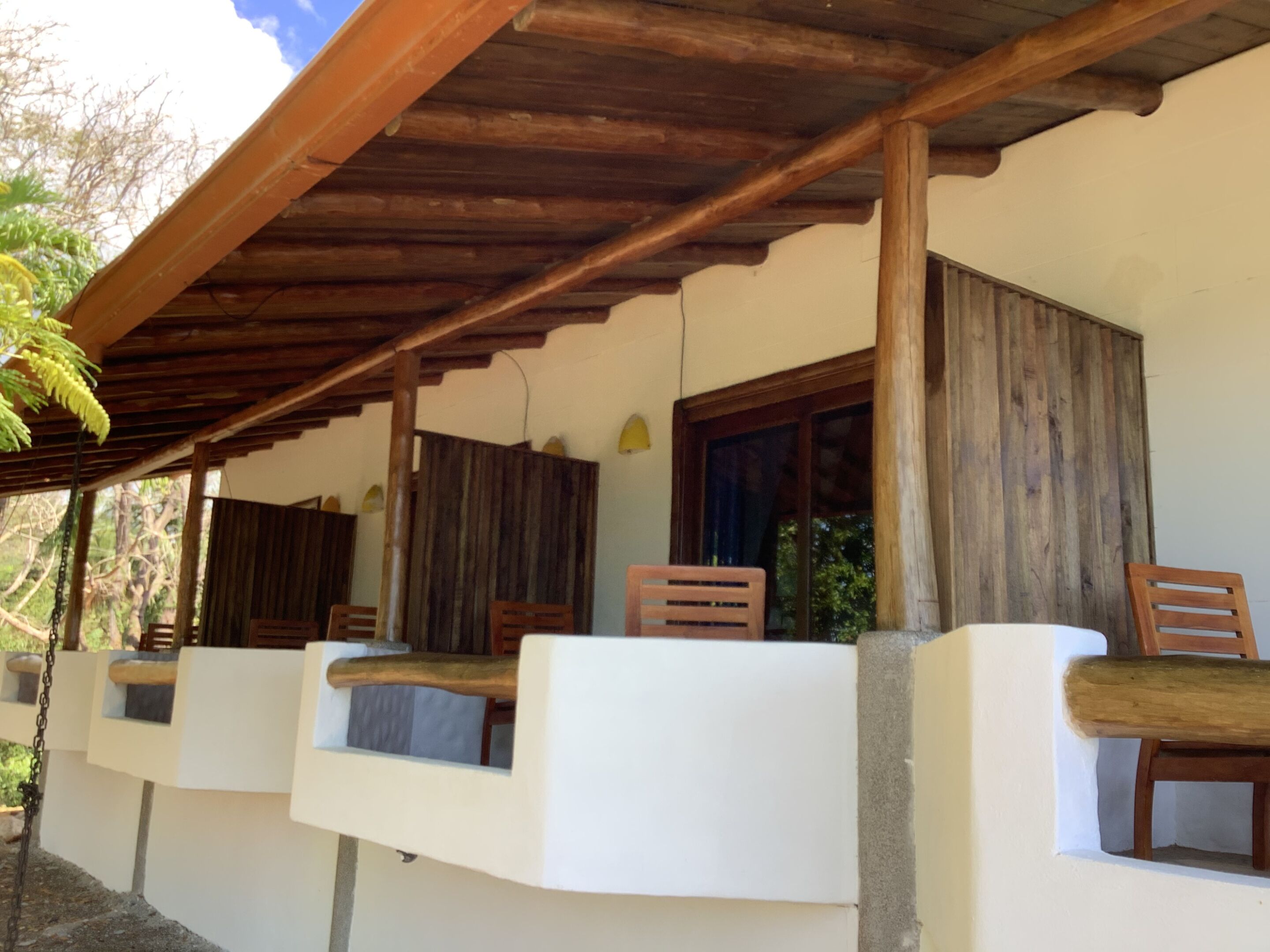 A covered outdoor seating area with wooden chairs and a white wall.