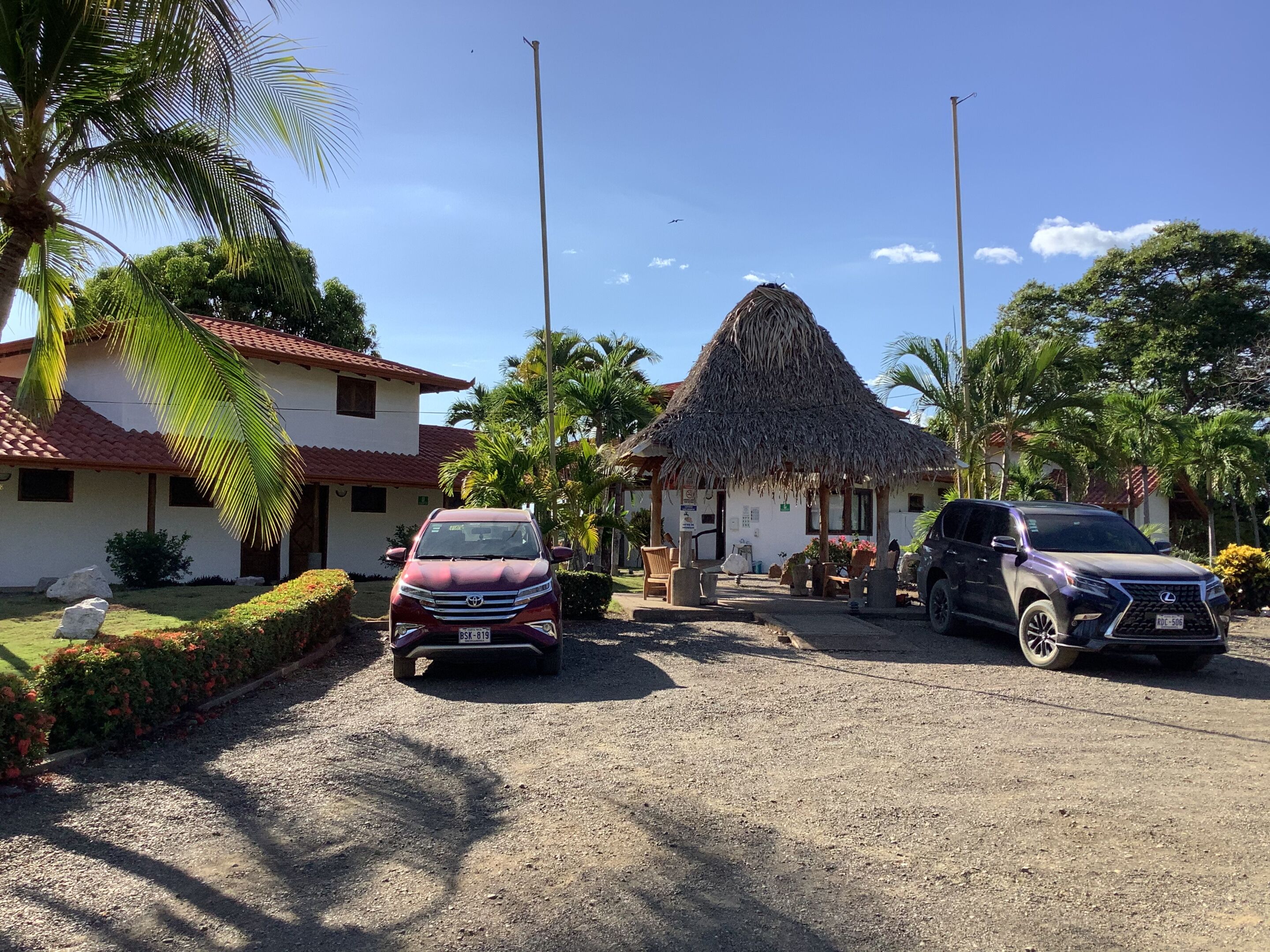 A hotel with a thatched-roof structure, a red car, and a black SUV parked in front.