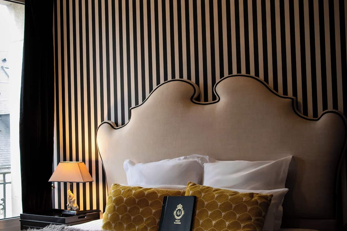 Une chambre d’hôtel avec un lit, un papier peint à rayures, un lustre et une table de chevet avec une lampe.