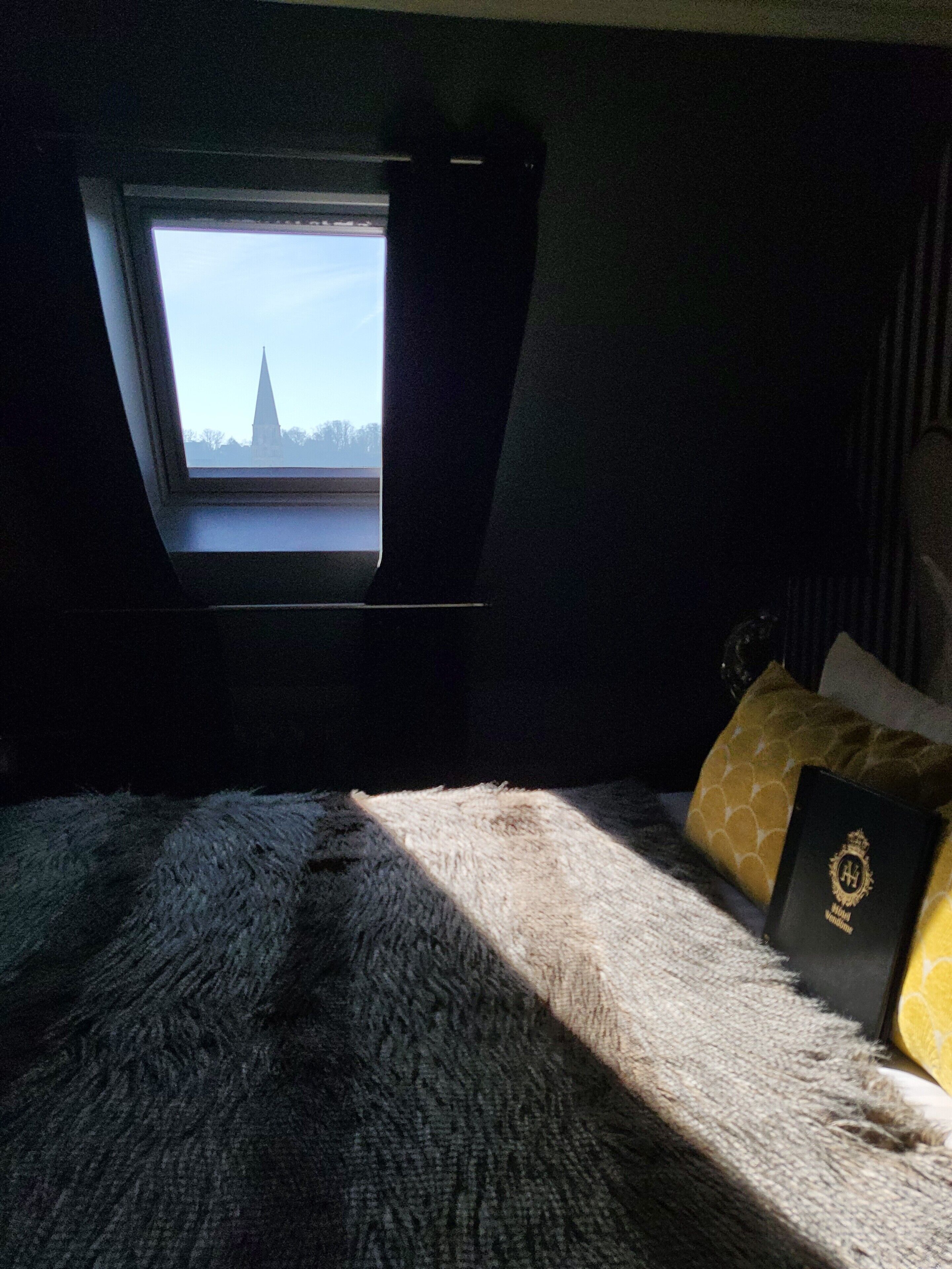 Un dormitorio con ventana que da a un paisaje urbano, una cama con una manta mullida y un almohadón con diseño dorado.