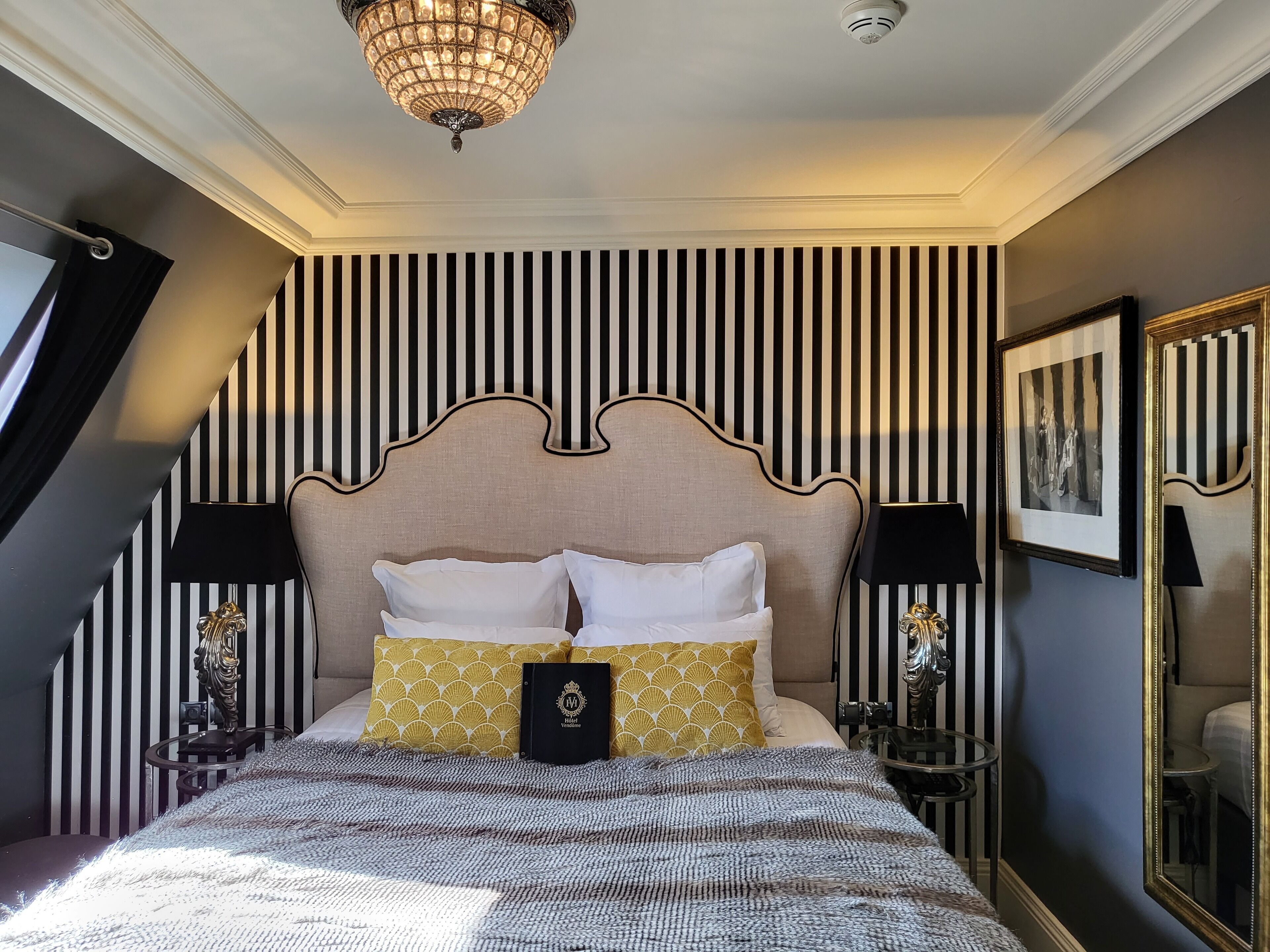 A hotel room with a bed, striped wallpaper, a chandelier, a mirror, and a framed picture on the wall.
