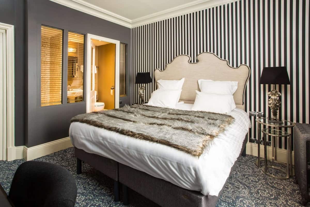 Une chambre d’hôtel avec un grand lit, une table de chevet avec une lampe, une chaise et une vue sur la salle de bain à travers une porte.