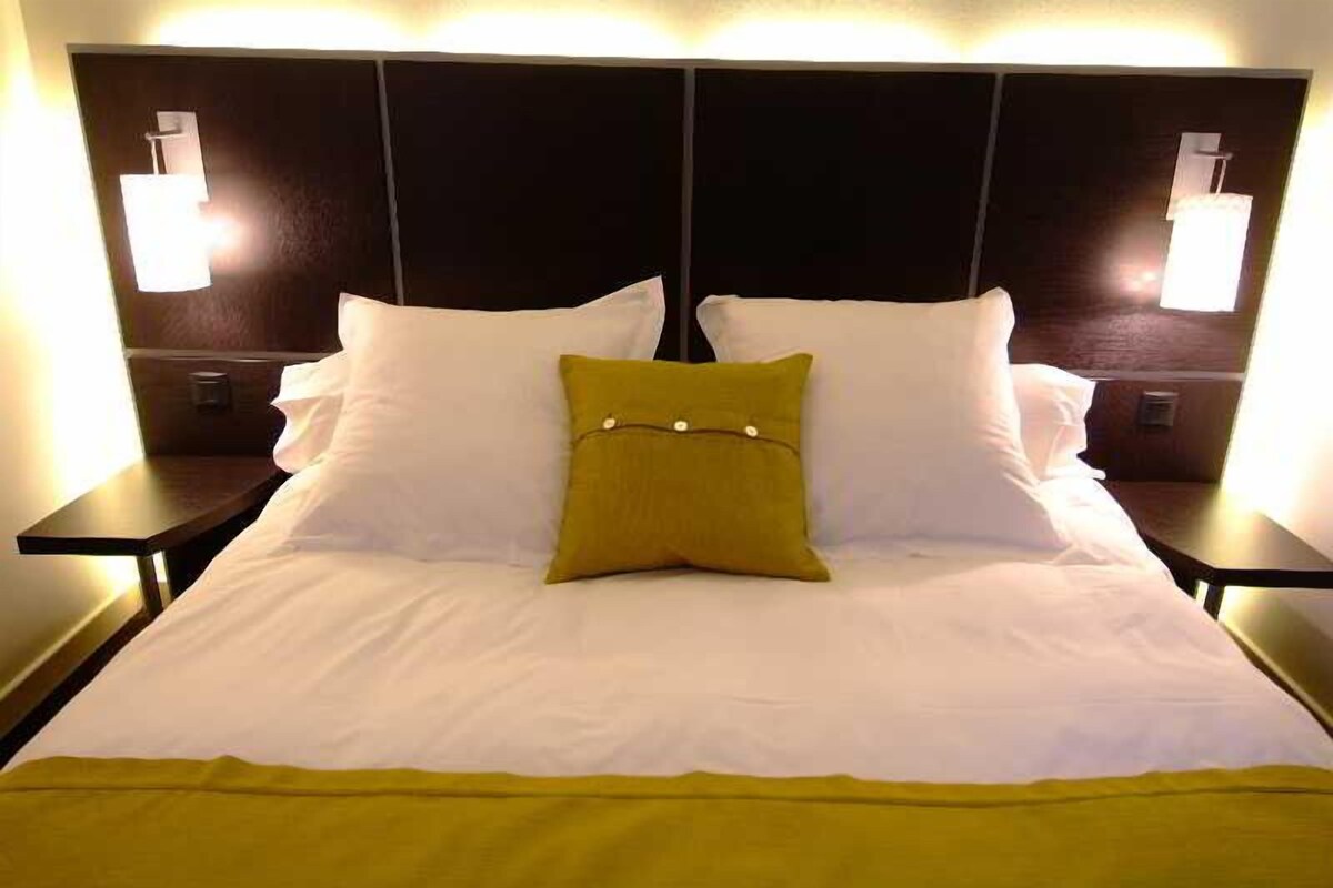 Une chambre d’hôtel avec un grand lit, deux tables de chevet et des lampes fixées au mur.