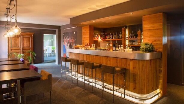 Zona de bar moderna con mostrador de madera, taburetes y una variedad de botellas y vasos.