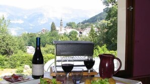A table set with plates of food, a bottle of wine, glasses, and a laptop on a balcony with a scenic view of a village and mountains. - Chalet Liberty Mont-blanc (Saint-Gervais-les-Bains)