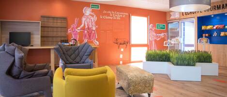 Un interior moderno con una zona de estar que incluye una silla amarilla, un sillón gris y un pequeño reposapiés. Hay un televisor sobre una estantería de madera, una pared con una ilustración de un cerdo, y un letrero que dice "La cité de Carcassonne, le Lochon et le Champ de ble".