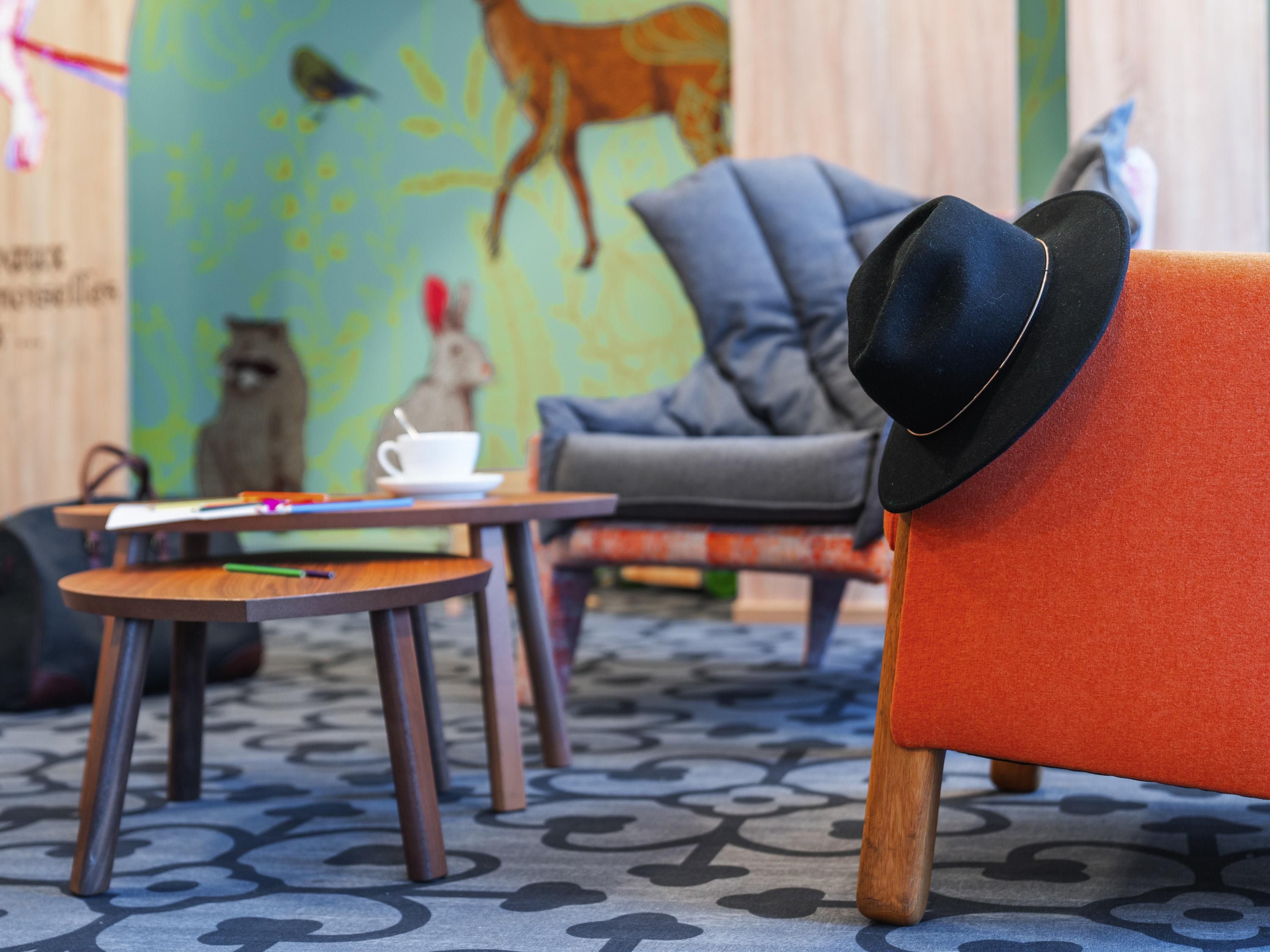 A room with a patterned floor, a grey sofa, a small round table with a cup and a book, and a red armchair with a black hat on it.