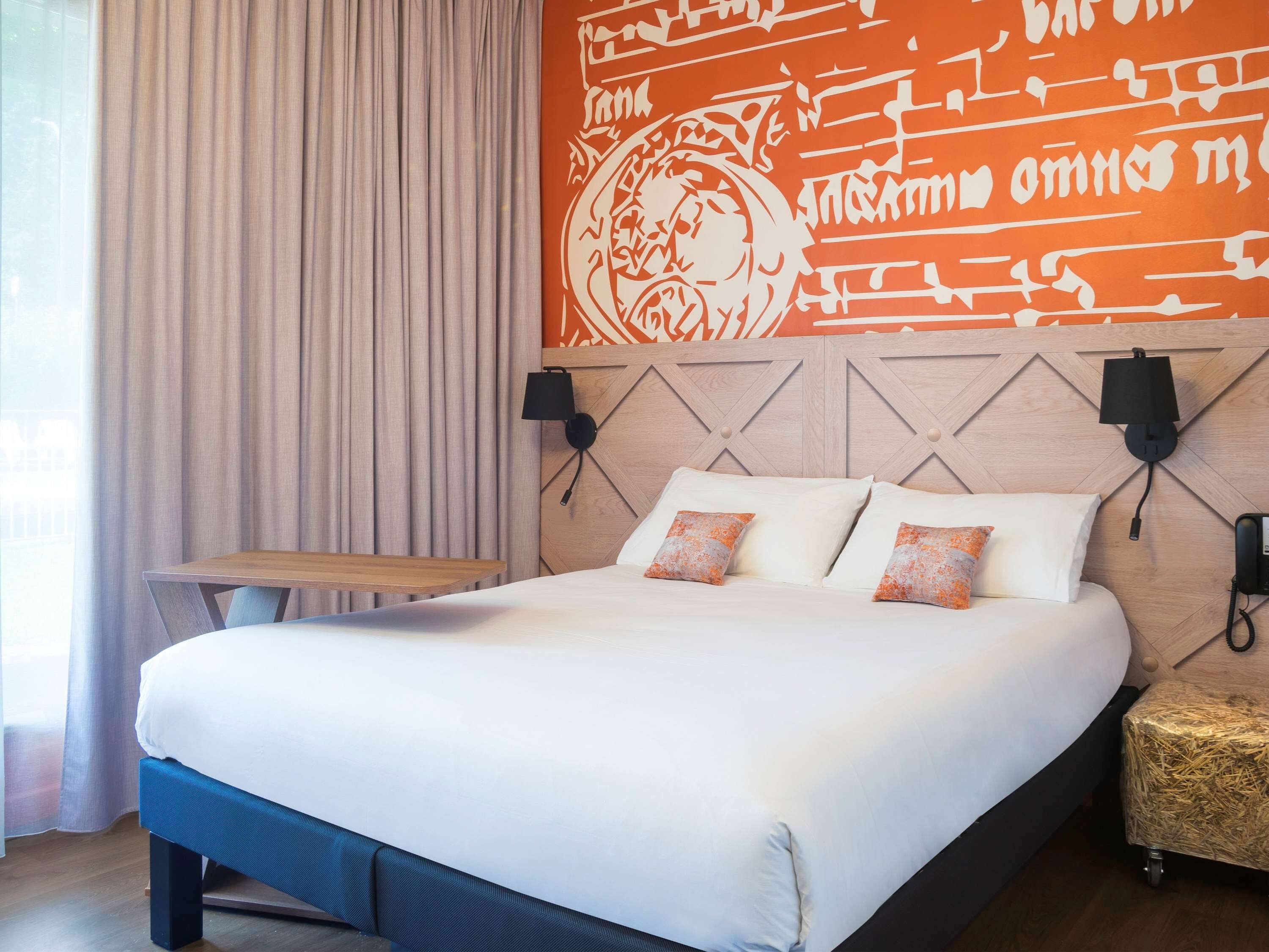 A hotel room with a large bed, a wooden headboard, two wall-mounted lamps, and a small table.