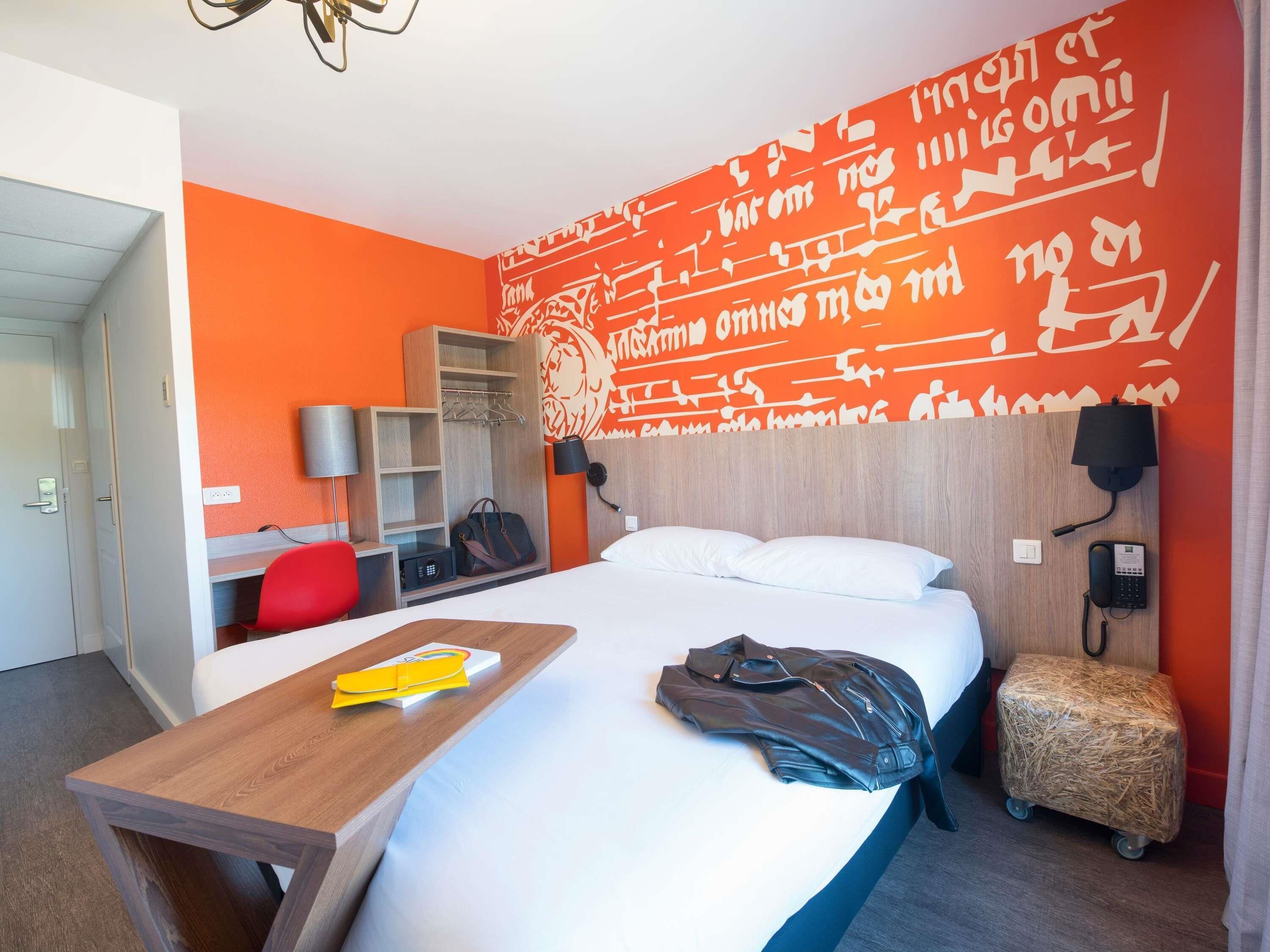 Une chambre d’hôtel avec un lit, un bureau en bois, une chaise rouge, une armoire et un mur orné d’une écriture décorative.