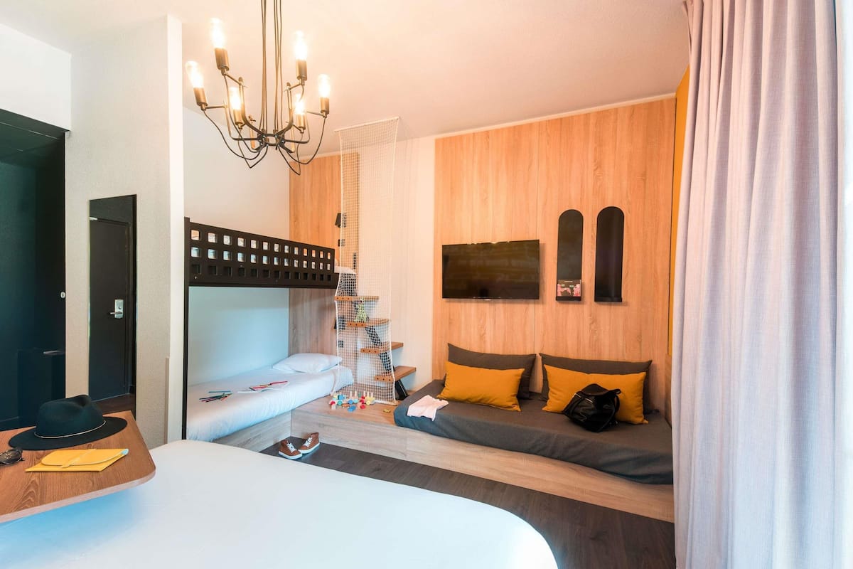 Quarto de hotel moderno com beliche, uma escrivaninha com cadeira, um lustre e uma televisão fixada na parede.