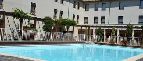 A swimming pool with a wooden deck, surrounded by a white fence and a building with multiple windows and balconies.