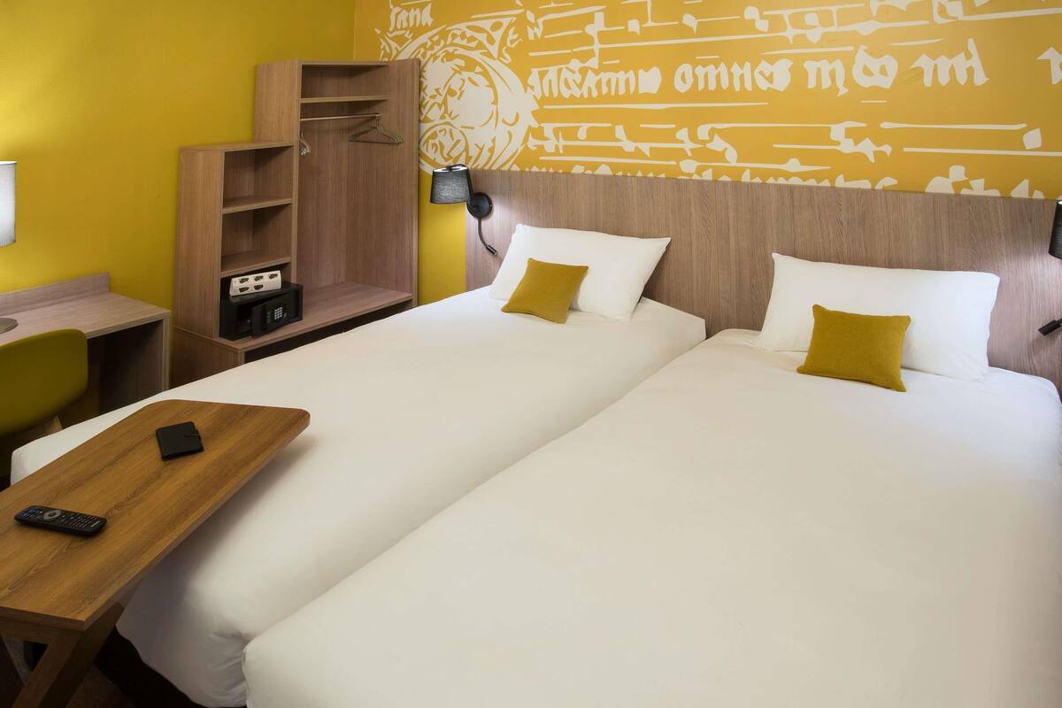 Quarto de hotel com duas camas, mesa de cabeceira de madeira, escrivaninha e parede com padrões decorativos.