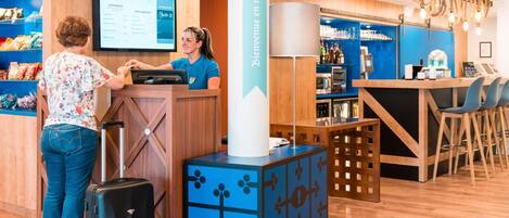 Mostrador de recepción de hotel con una pantalla digital que muestra tarifas de habitaciones, una zona de bar con taburetes y un área de almacenamiento de equipaje.