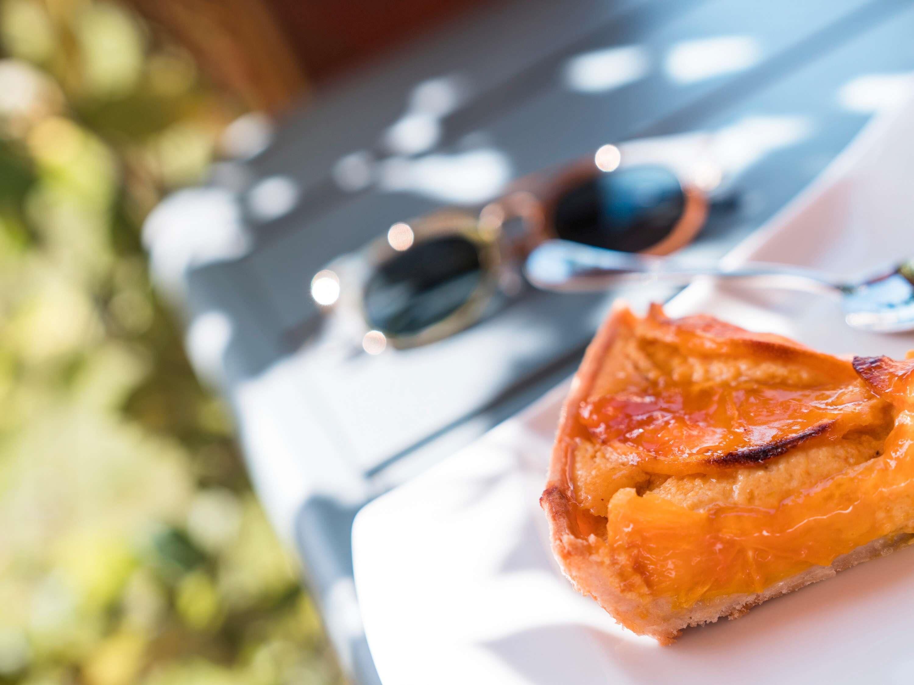 Una porción de tarta de crema de naranja sobre un plato blanco, con un par de gafas de sol y un fondo exterior desenfocado.