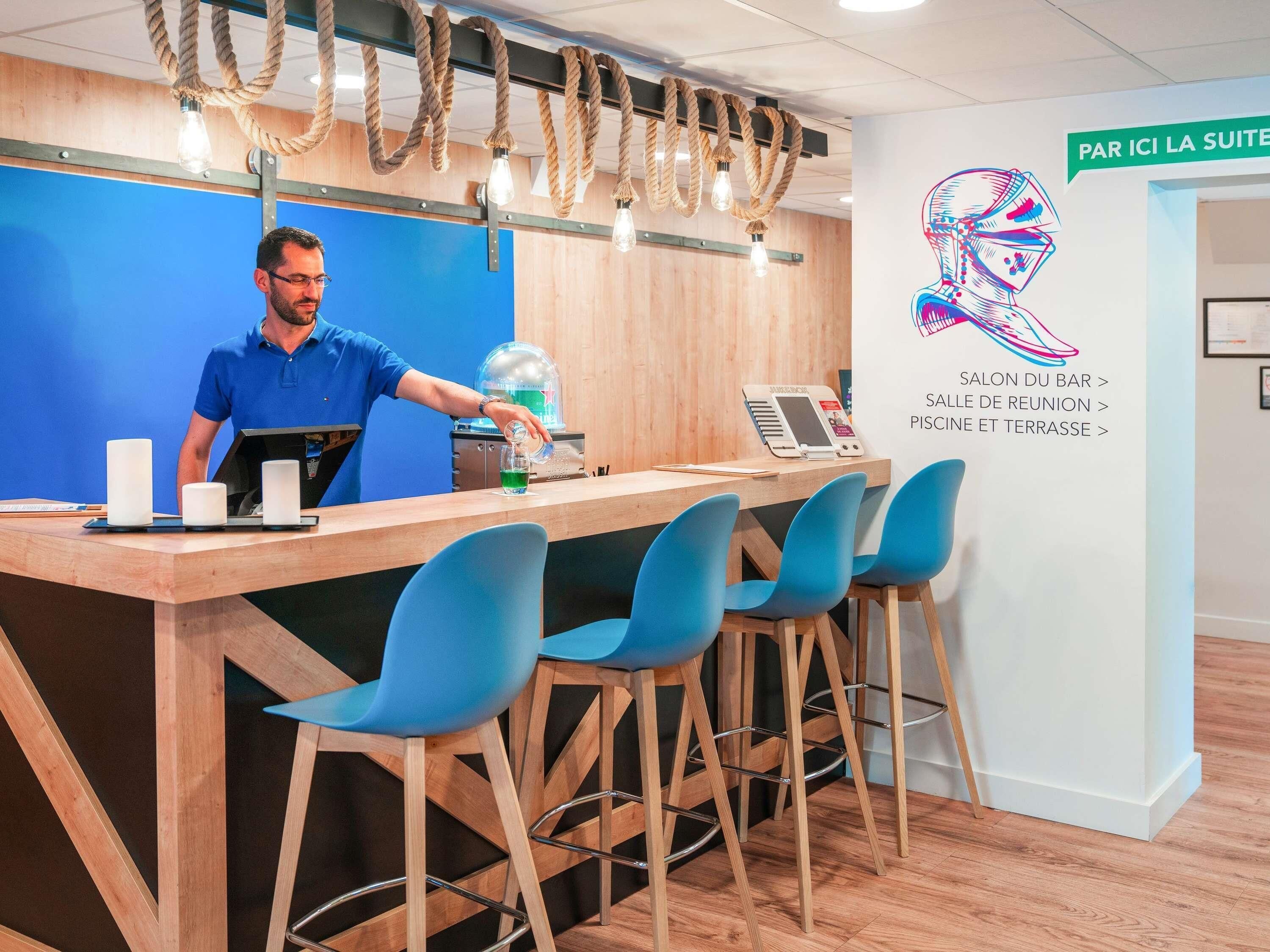 A reception desk with a man standing behind it, a bar area with blue chairs, and a wall with directional signs.
