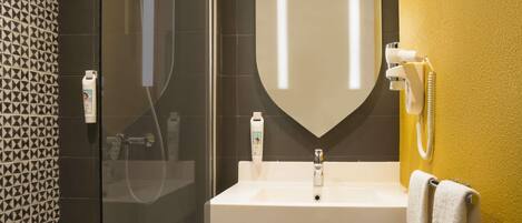 Une salle de bain dotée d’une douche à parois de verre, d’un lavabo blanc et d’un miroir au design évoquant un bouclier.