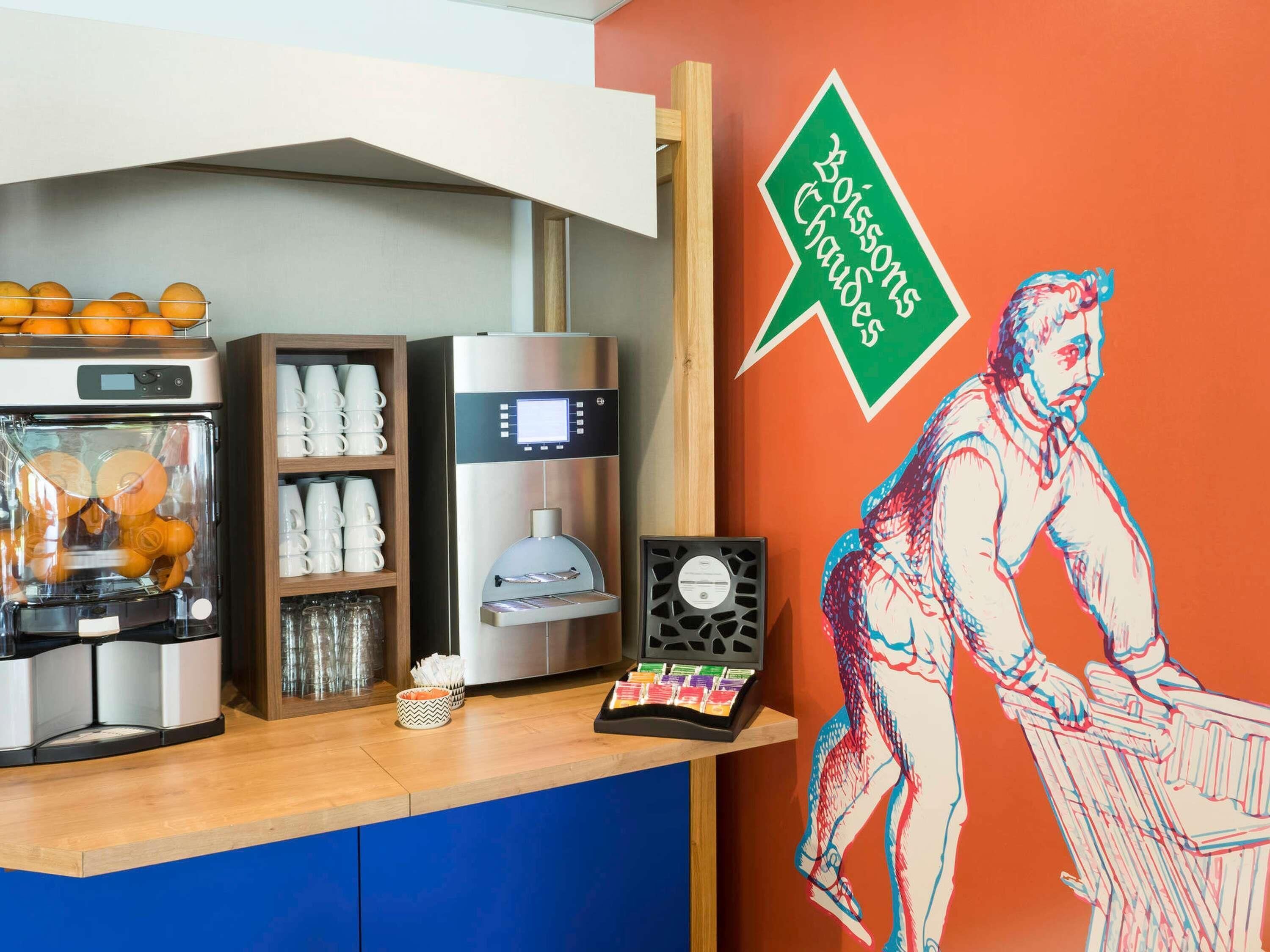 A coffee station with a coffee machine, a glass display case with oranges, and a wall with a green sign and a pop art painting.