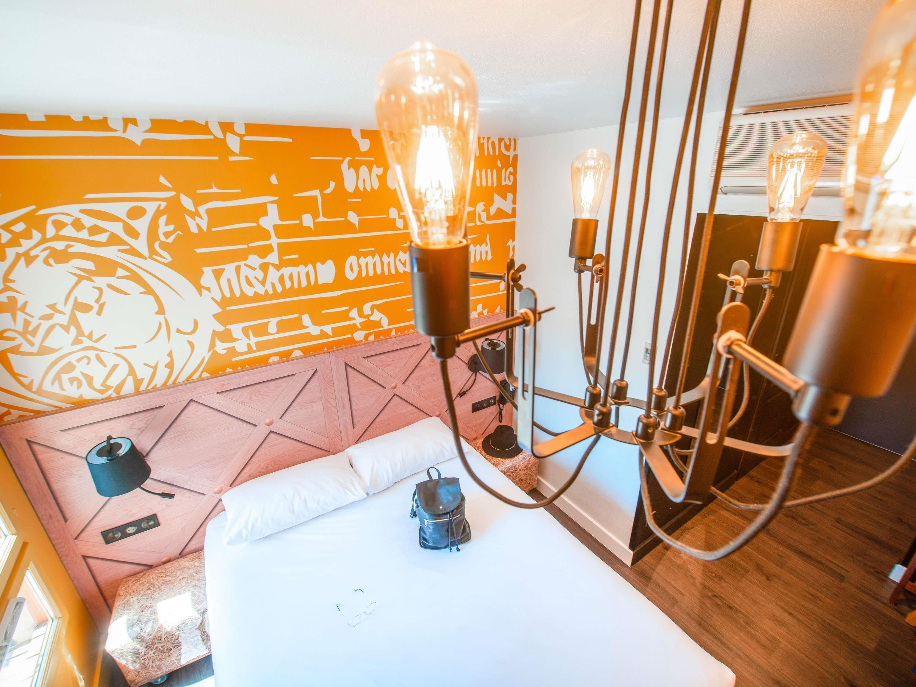 Habitación de hotel con cama, pared con diseño geométrico y candelabro con varias bombillas.