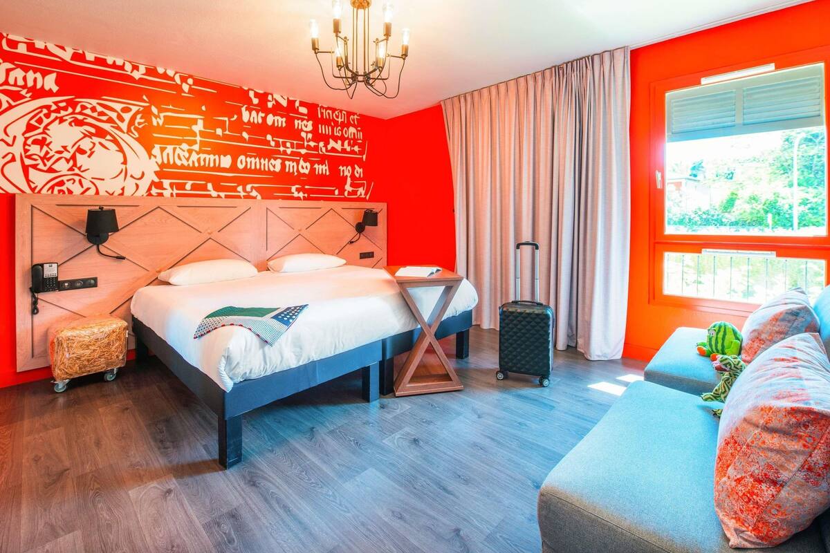 Quarto de hotel moderno com cama, sofá, mala e janela com cortinas.
