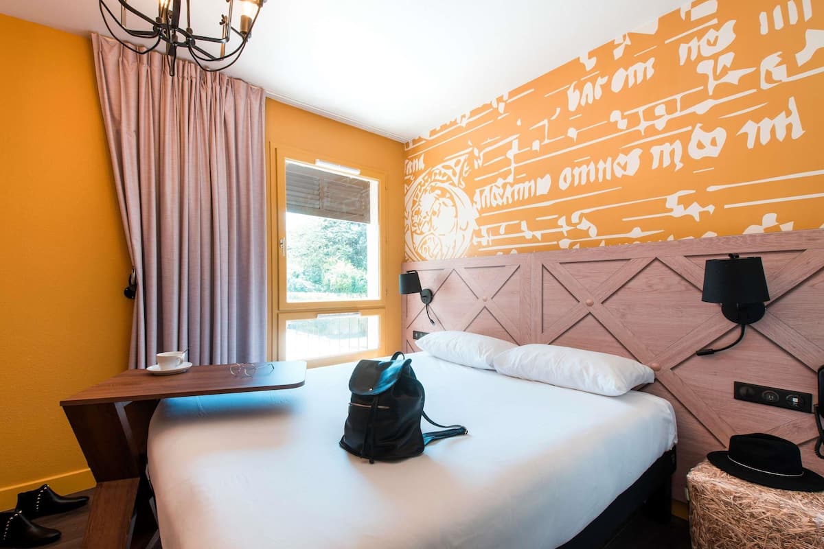 Quarto de hotel com uma cama, escrivaninha, cadeira, mala e uma pequena mesa.