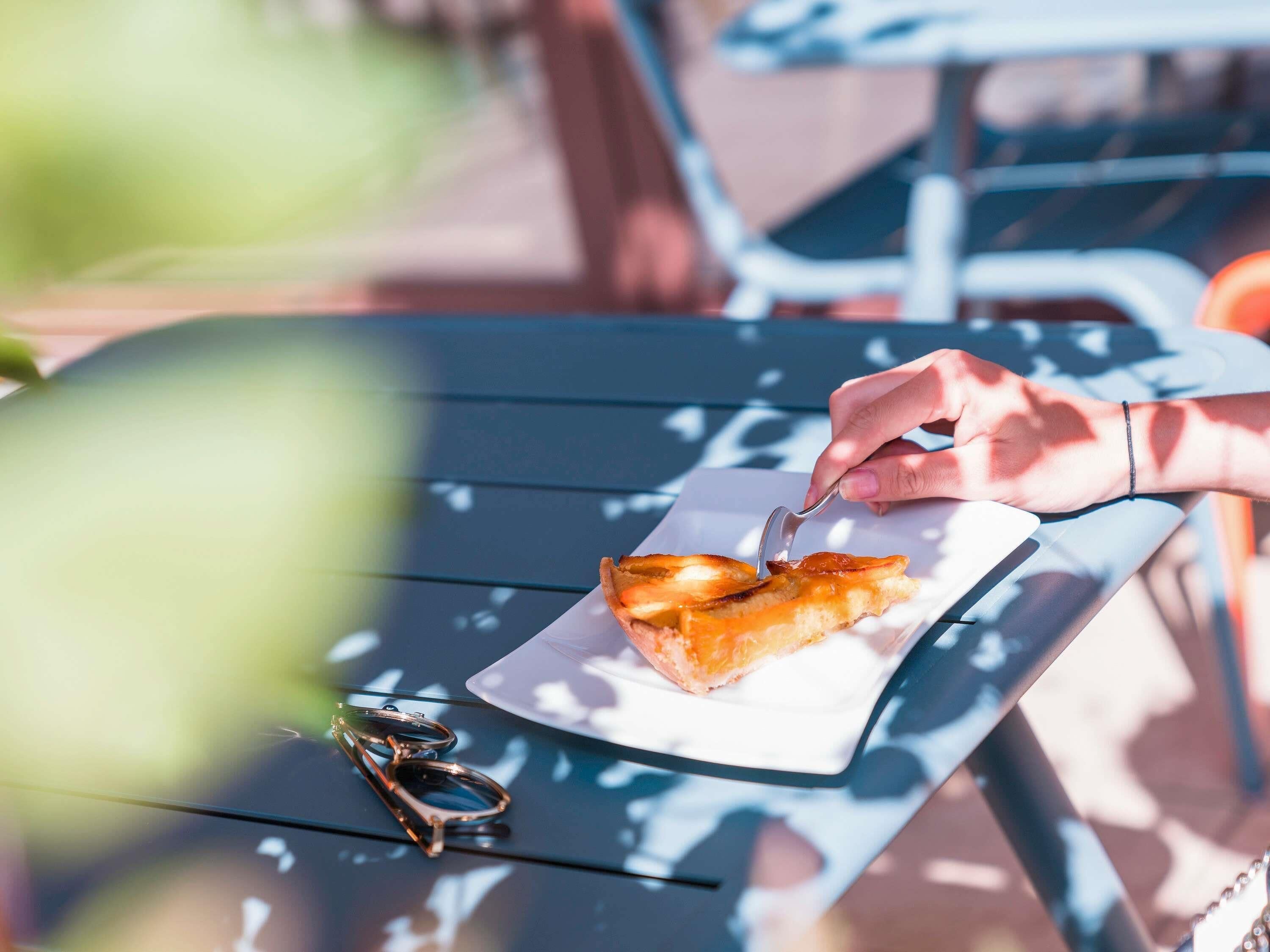 A hand holding a spoon over a plate with a piece of food, a pair of sunglasses, and a table with a metal frame.