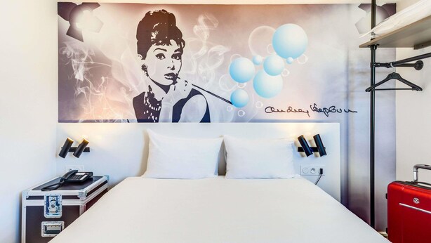 Une chambre d’hôtel avec un lit, une grande fresque murale, une valise rouge et une valise noire.