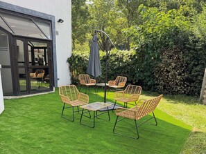 Une terrasse avec quatre chaises en osier et une table, entourée d’une haie et d’un bâtiment blanc doté d’une porte en verre.