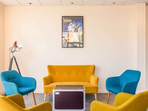 Una habitación con un sofá amarillo, dos sillones azules y una pequeña mesa. Hay un cartel en la pared y un trípode con una lámpara en el suelo.