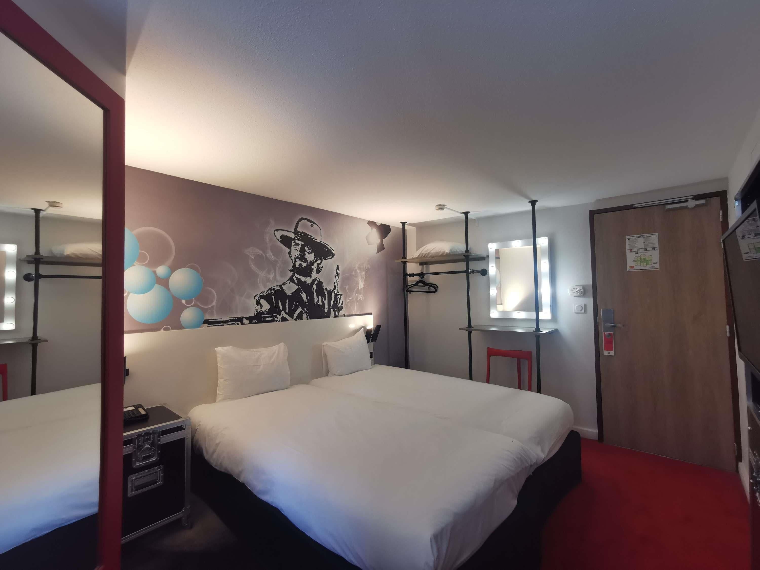 Een hotelkamer met een groot bed, een spiegel met verlichting, een rode stoel en een muur met een afbeelding.