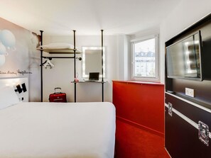 Habitación de hotel con cama, pared decorada en rojo, espejo grande, ventana con vistas a la ciudad y televisor.