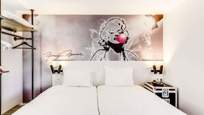 A hotel room with a large bed, a wardrobe, a nightstand with a phone, and a wall with a signature and a pop art-style painting.
