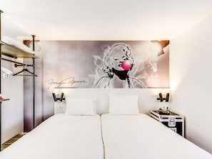 Een hotelkamer met een groot bed, een kledingkast, een nachtkastje met een telefoon, en een muur met een handtekening en een popart schilderij.