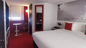 A hotel room with a bed, a desk, a chair, a mirror, a nightstand, and a wall with a signature.