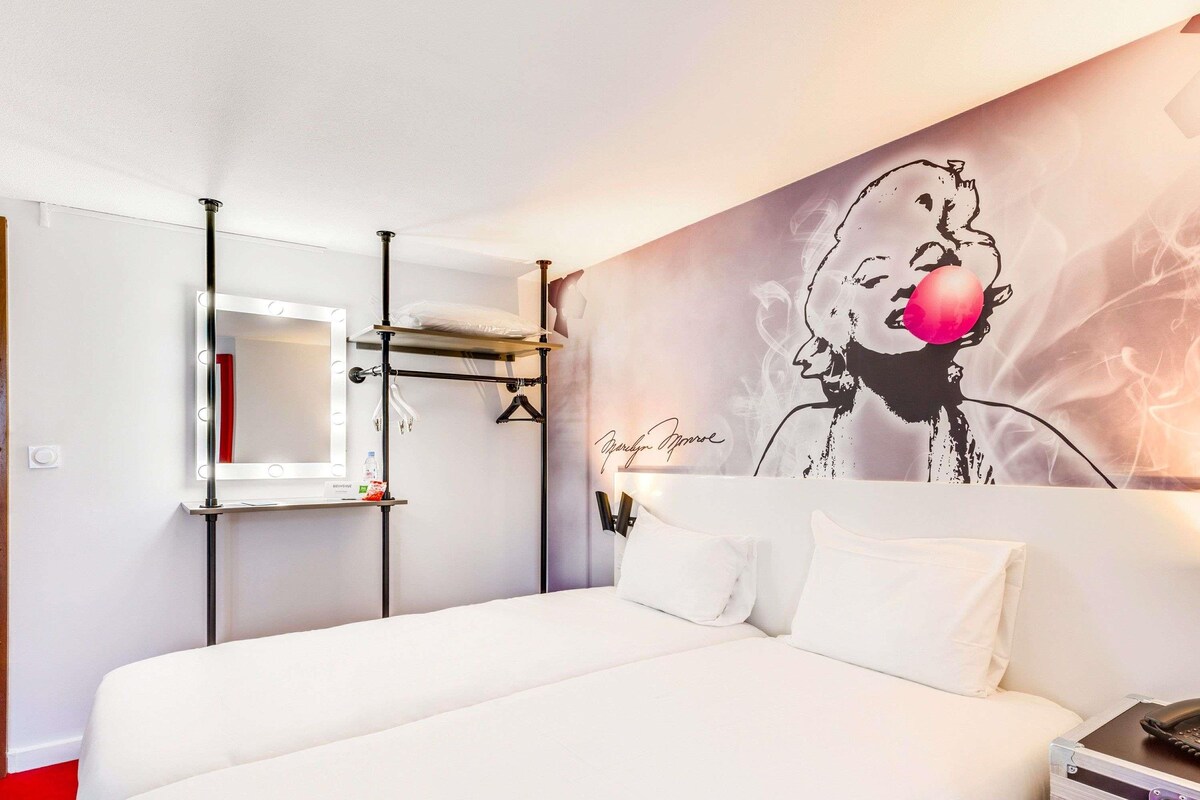 Une chambre d’hôtel avec un lit, une coiffeuse dotée d’un miroir et de lampes, un mur orné d’une fresque, et une valise posée sur une table.