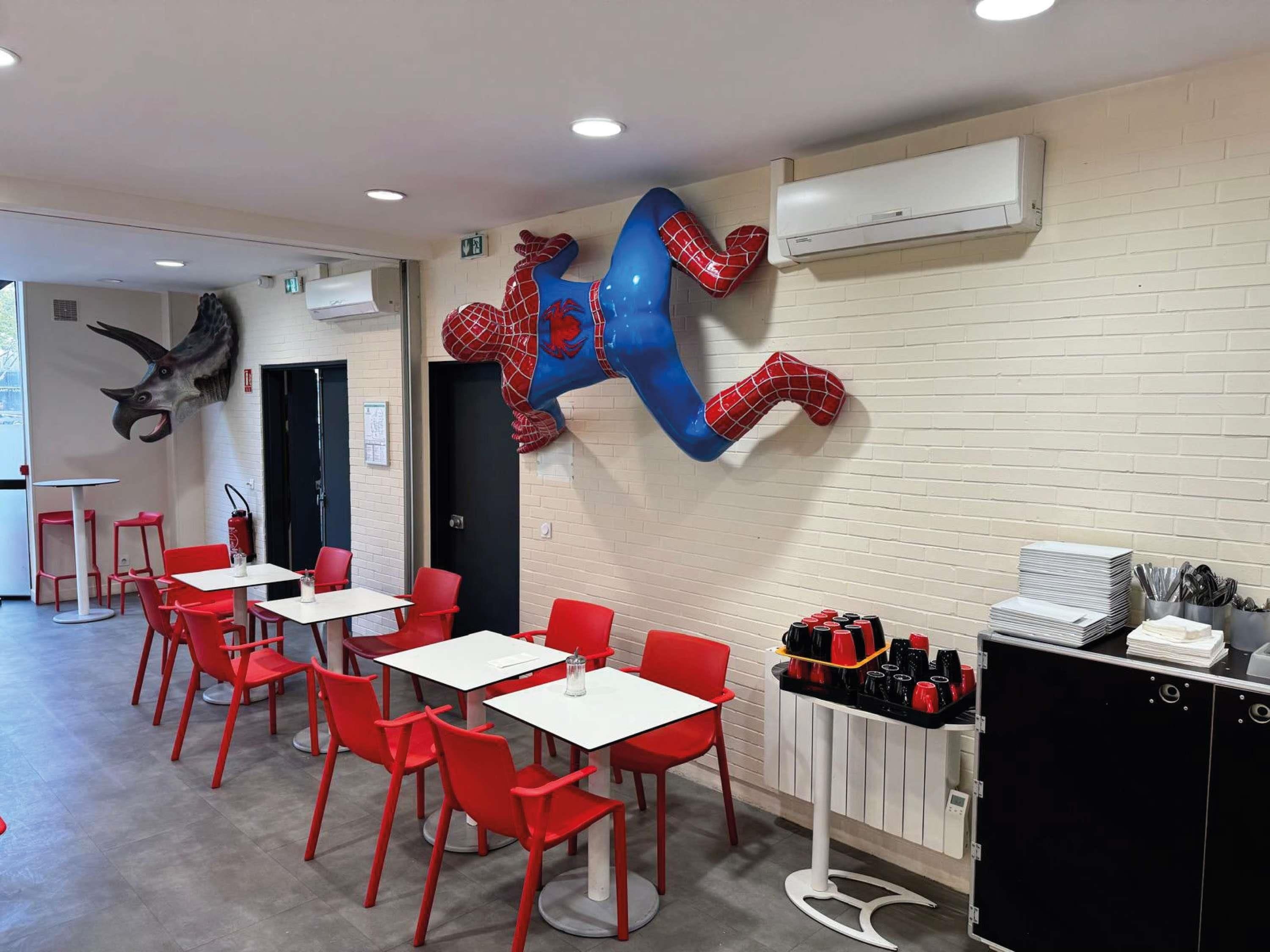 Un espace repas avec des chaises rouges et des tables blanches, une sculpture de Spiderman fixée au mur et un climatiseur mural.