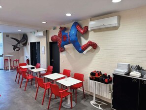 Un comedor con sillas rojas y mesas blancas, una escultura de Spiderman montada en la pared y un aire acondicionado instalado.