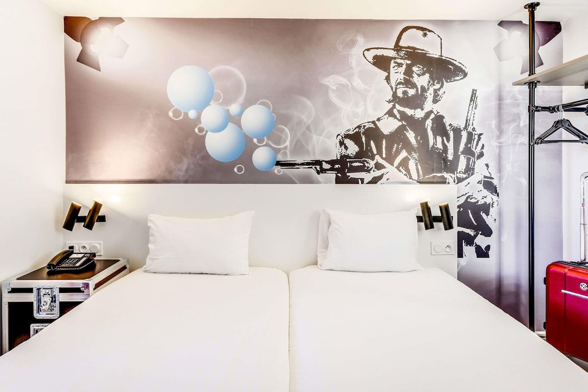 Hotelkamer met een groot bed, twee kussens, een nachtkastje met een telefoon en een lamp, en een muur waarop een afbeelding te zien is van een man met een pistool en ballonnen.