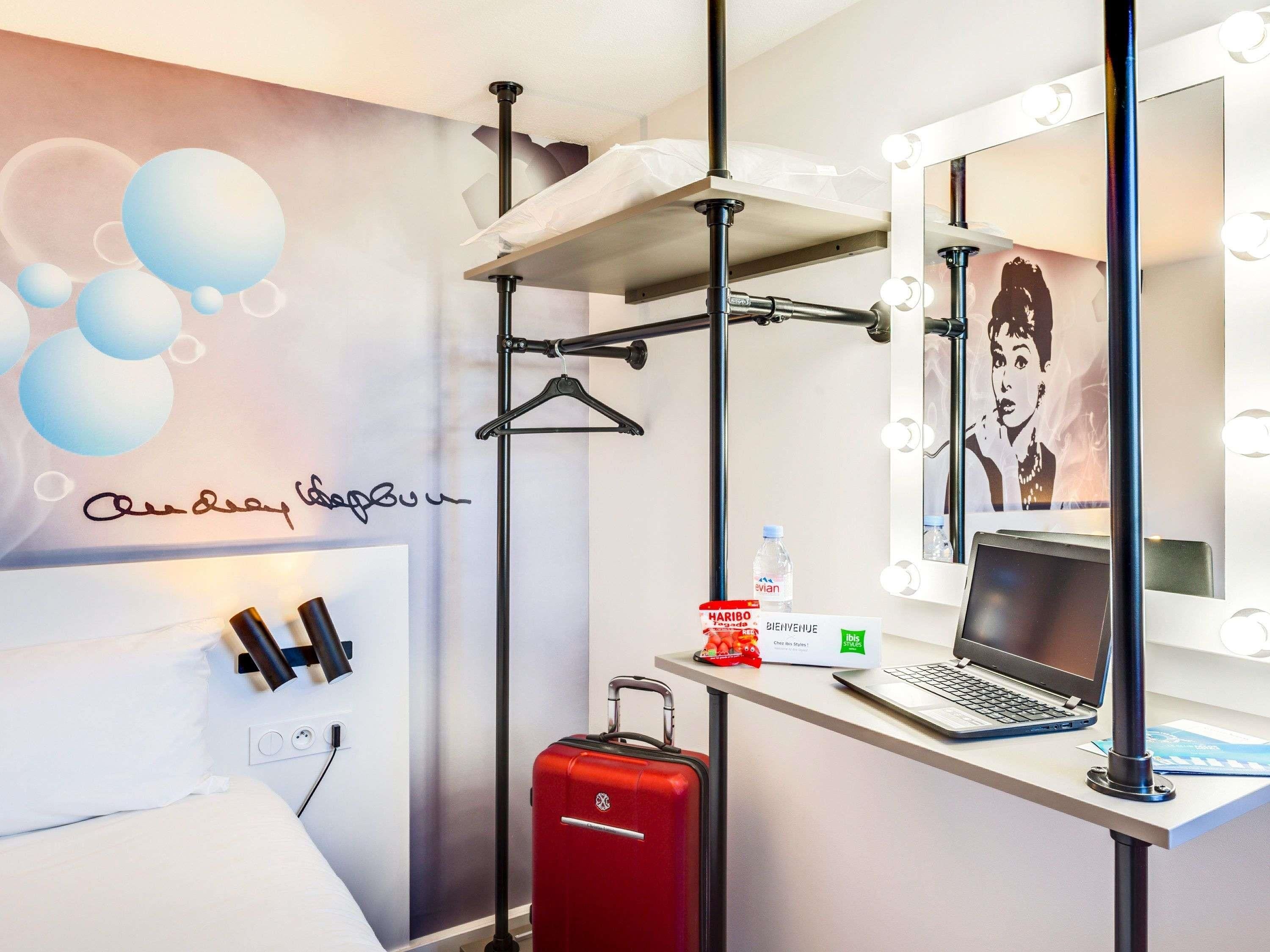 A hotel room with a bed, a red suitcase, a desk with a laptop, a mirror with lights, and a wall with a signature.