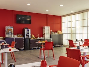 Un espace repas moderne avec des chaises et des tables rouges, une machine à café et un téléviseur fixé au mur.