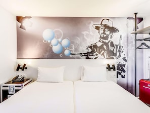 A hotel room with a large bed, two pillows, a bedside table with a phone and a lamp, and a wall with a graphic of a man with a gun and balloons. - ibis Styles Paris Saint Denis Plaine (Saint-Denis)