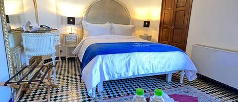 Habitación de hotel con suelo a cuadros, cama con colcha blanca y azul, mesa de centro redonda con dos botellas de agua, espejo grande y armario de madera.